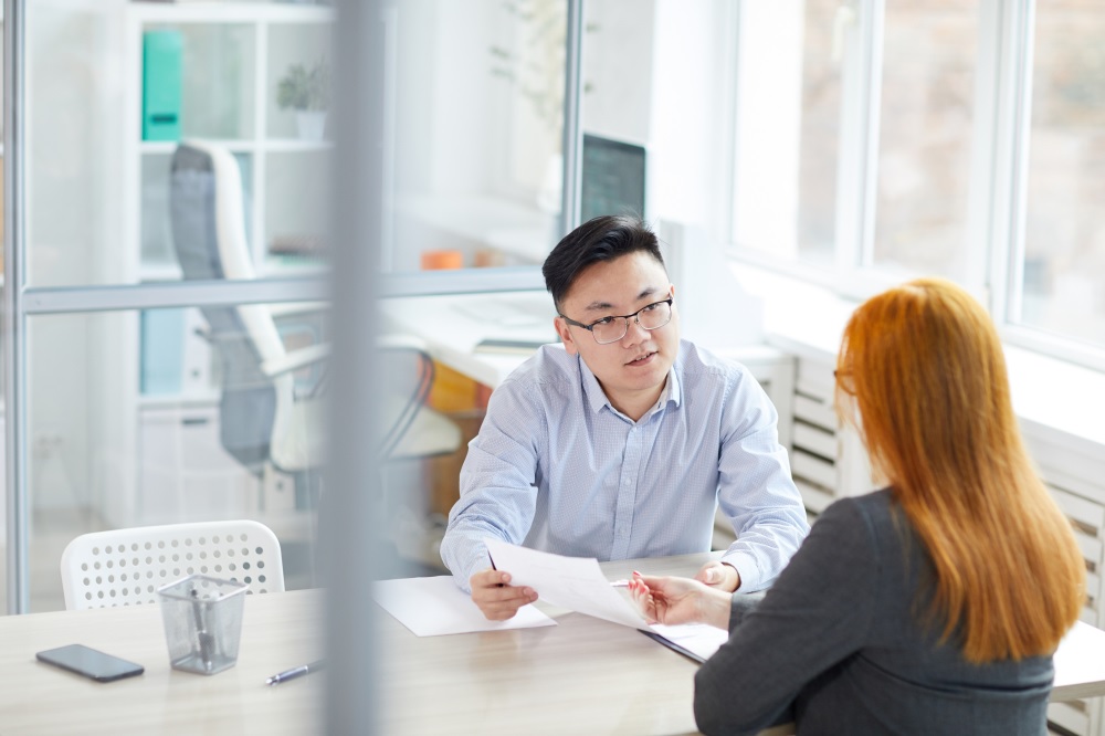 Image shows an office meeting rooms where a man, who is facing the camera, is talking to a lady. They represent using one of the sSix effective recruitment strategies to successful hiring.